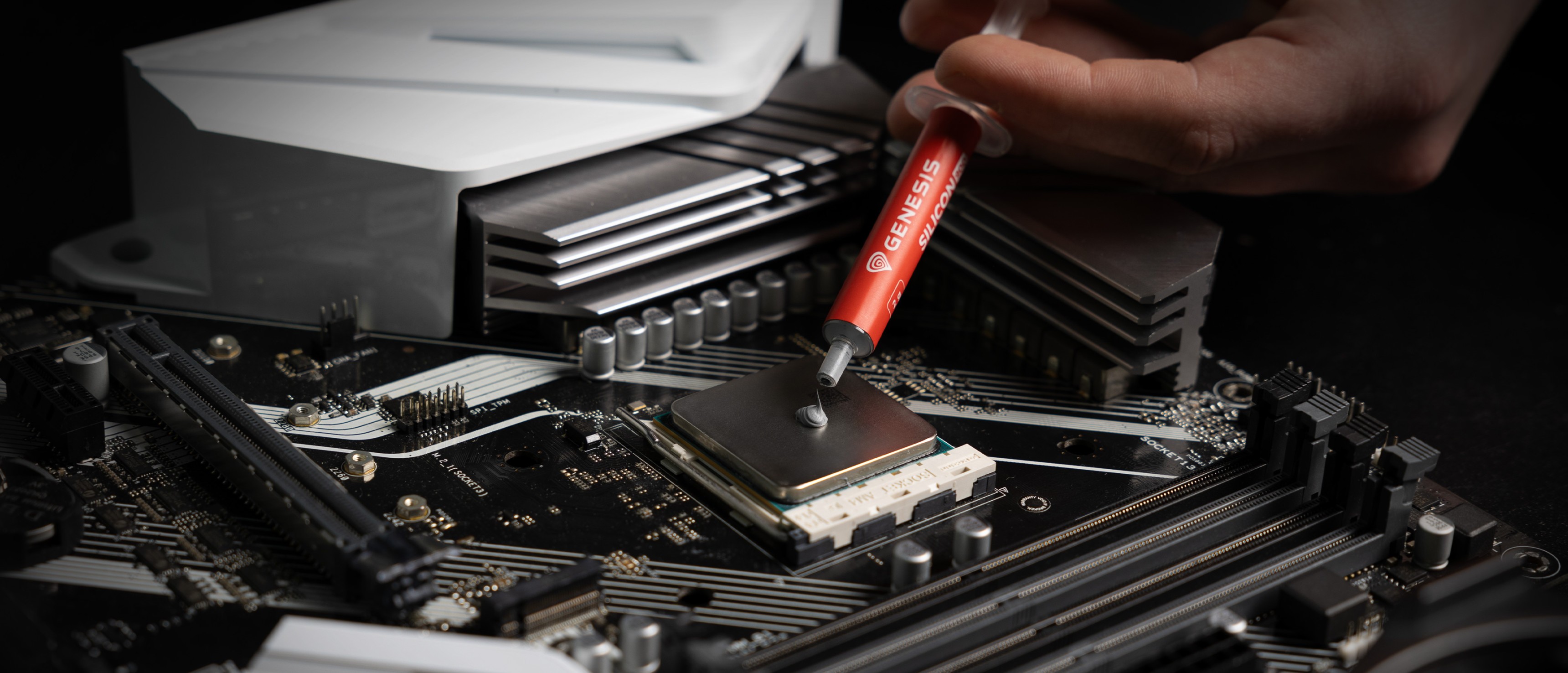Wärmeleitpaste wird aus einer Spritze auf einen CPU-Sockel auf einem Motherboard aufgetragen, wobei Kühlkomponenten sichtbar sind