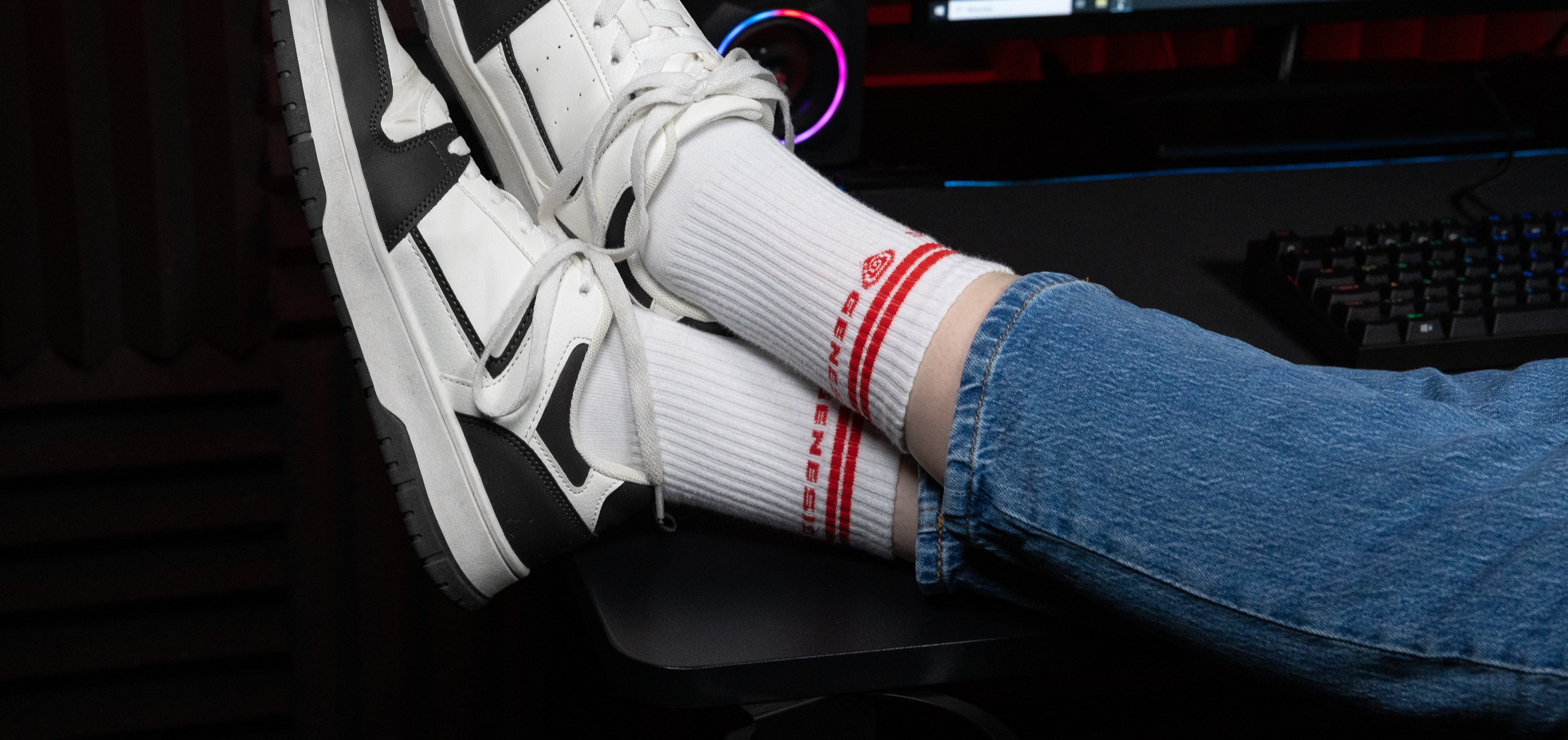 Weiße Sportsocken mit rotem Branding, getragen zu weißen und schwarzen Turnschuhen, auf einem Schreibtisch mit einem Computer-Setup liegend